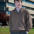 A man with hands in his pockets stands smiling in front of a large concrete building.