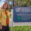A blonde woman with glasses, wearing a yellow shirt and a motley scarf, stands smiling on a sidewalk in front of trees beside a sign marking “UF University IFAS Extension State Headquarters Florida 4-H Youth Development.”