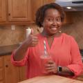 A lady in a kitchen holds a smoothie and gives a thumbs up.