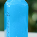 Close-up of a portion of a clear bottle filled with bright blue liquid that is water-soluble fertilizer dissolved in water, against a blurred dark green background.