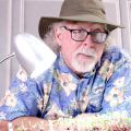 A man with a white goatee and dark-framed glasses leans on a table behind a small LED table lamp and a tray of seedlings. He wears a blue floral Hawaiian shirt and brown hat. 