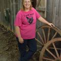 Dressed in a pink T-shirt and blue jeans, broiler grower Teresa Dyess stands next to two wagon wheels in front of a barn on her family farm.