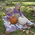Two young girls sit on a colorful quilt among leaves in the grass as they play with a white and an orange pumpkin.