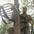 Hunter wearing camouflage secures a portable platform to the side of a tree.