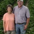 A wife and husband stand in a field with cotton rows.