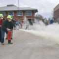 Two men wearing hard hats and masks activate a fire extinguisher as MyPI training participants watch. 