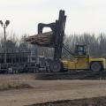 A yellow piece of heavy machinery lifts a load of cut trees off the back of a log truck in a sawmill yard.