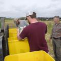 Two young men pour seed into bright yellow bins while a man watches.