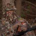 Only the eyes of a turkey hunter wearing full camouflage is visible. He is holding a wooden turkey caller.
