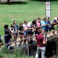 More than a dozen young people listen to a teacher while they stand on a cement bridge overlooking a large ditch.
