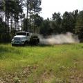 Dust billows out of a trailer on a large truck driving across a small, grassy area surrounded by tall trees.