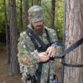 Man standing in the woods inspects nylon straps on a tree stand he is holding on in his hands.