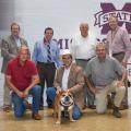 Seven men stand or kneel indoors in two rows, holding a bulldog in the center of the group.