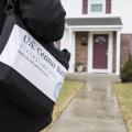 A person carrying a black bag marked with census information approaches the front door of a house.