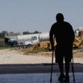 A man stands on crutches in silhouette against a background of farm equipment.