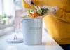 A woman scrapes vegetable scraps off a cutting board into a countertop compost bin.